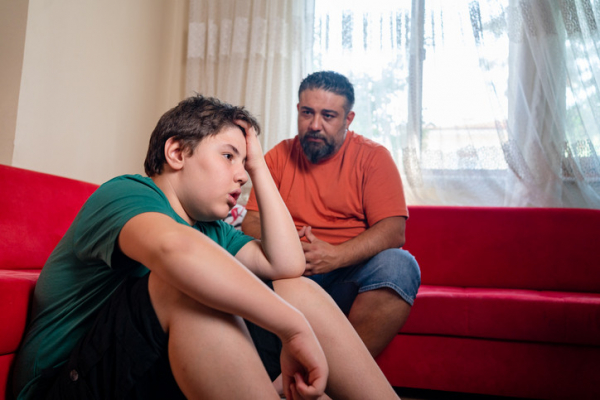 Bearded father with dark hair and orange shirt sitting on red couch; upset son in green shirt seated on floor nearby