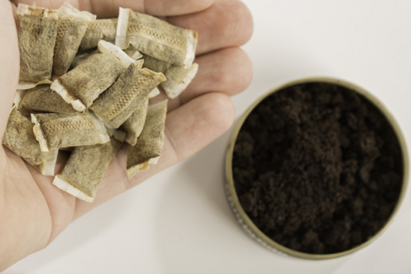 An open tin of dark brown smokeless tobacco known as snuff on right; fingers of a hand cupping pouches of snuff on left 